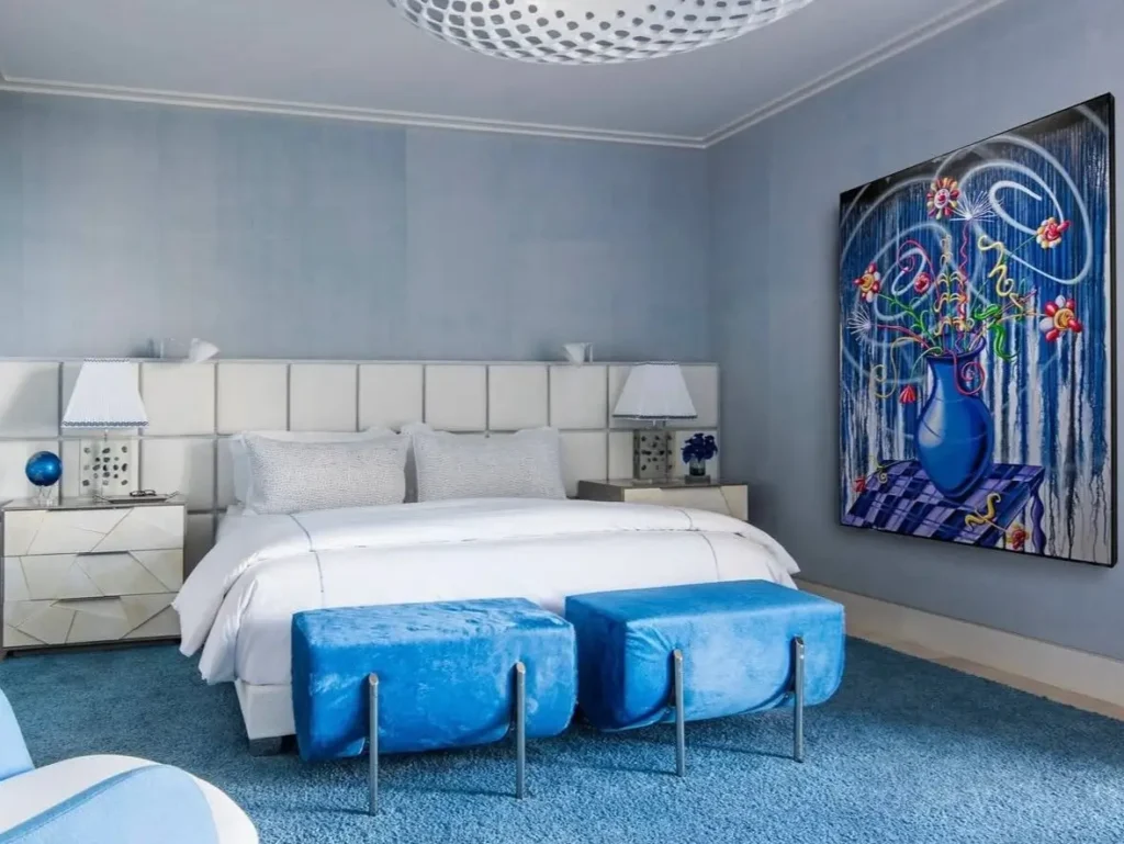 A modern beach themed bedroom with layered blue tones, a white upholstered headboard, and vibrant abstract art.
