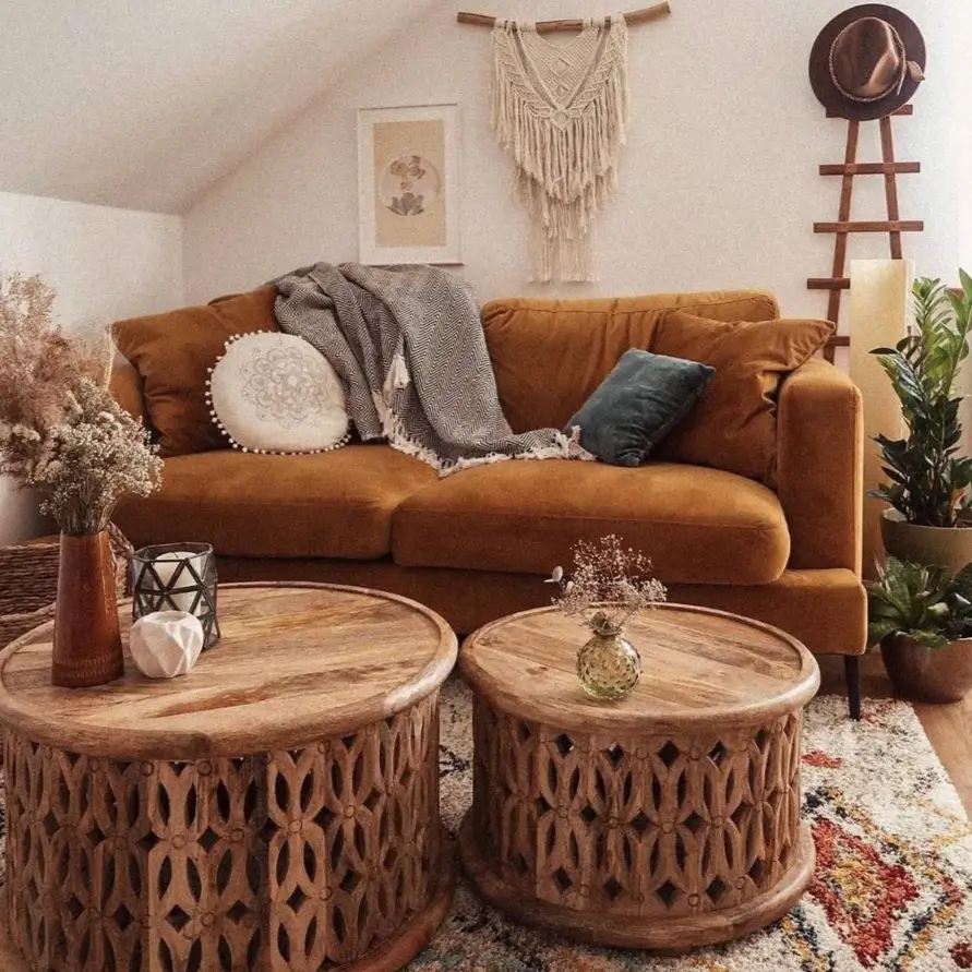 A bohemian living room with a mustard yellow sofa, two round carved wooden coffee tables, and a macrame wall hanging.