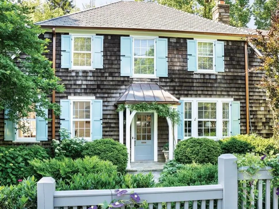 A cottage with natural cedar shingle siding, light blue shutters, and a small portico over the front door.