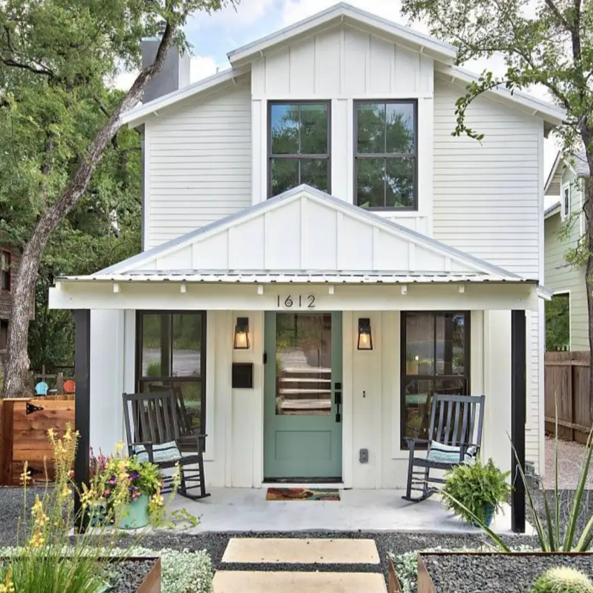 A modern white farmhouse cottage with a metal porch roof, black posts, a sage green door, and gravel landscaping.
