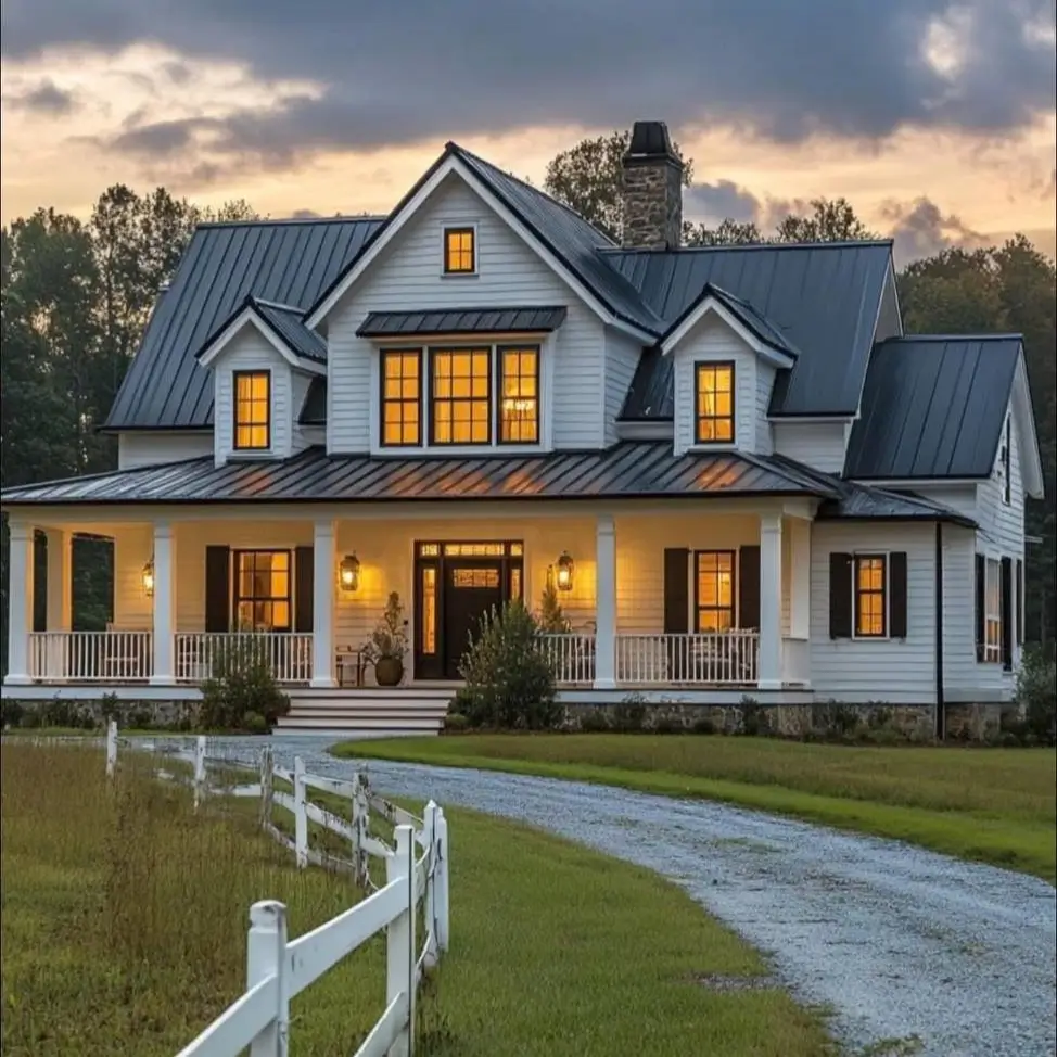 A modern white farmhouse with a dark metal roof and black shutters, glowing with warm lights at dusk.