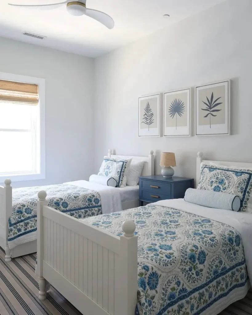 A coastal twin bedroom with white beds, blue floral bedding, and a series of three botanical prints on the wall.