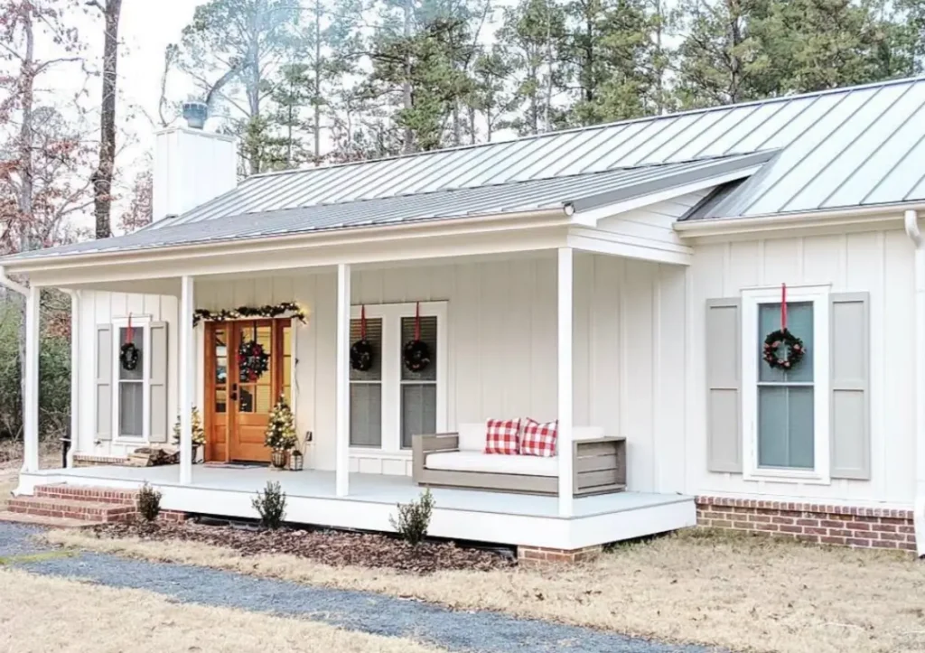 A white modern farmhouse cottage with a metal roof, a long front porch, and a natural wood door decorated with wreaths.