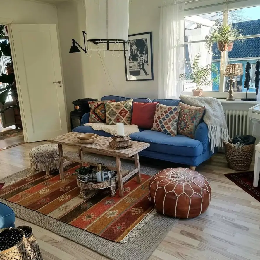 A bohemian living room with a blue sofa, a colorful striped rug, and a brown leather pouf in a sunlit room.