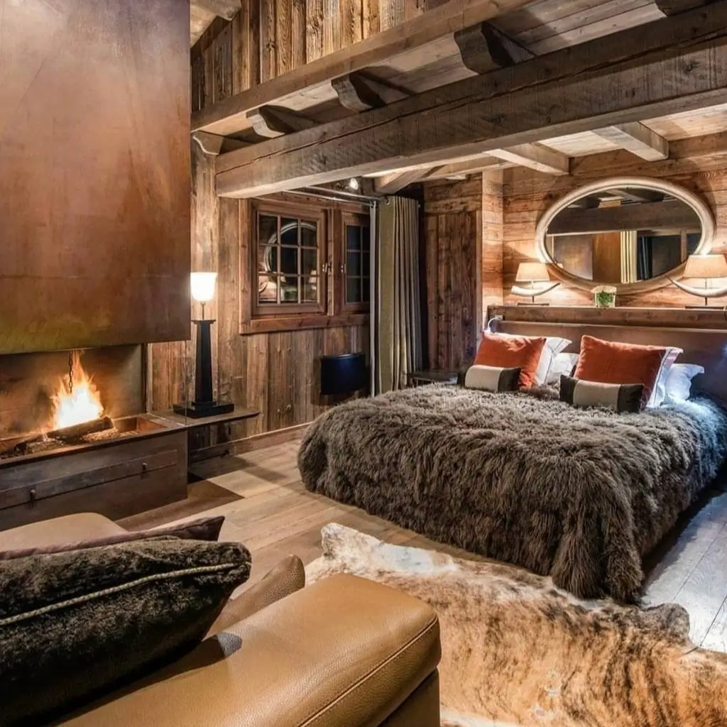 A cozy rustic bedroom for couples with a large fireplace, a bed covered in a dark fur throw, and exposed wooden beams on the ceiling.