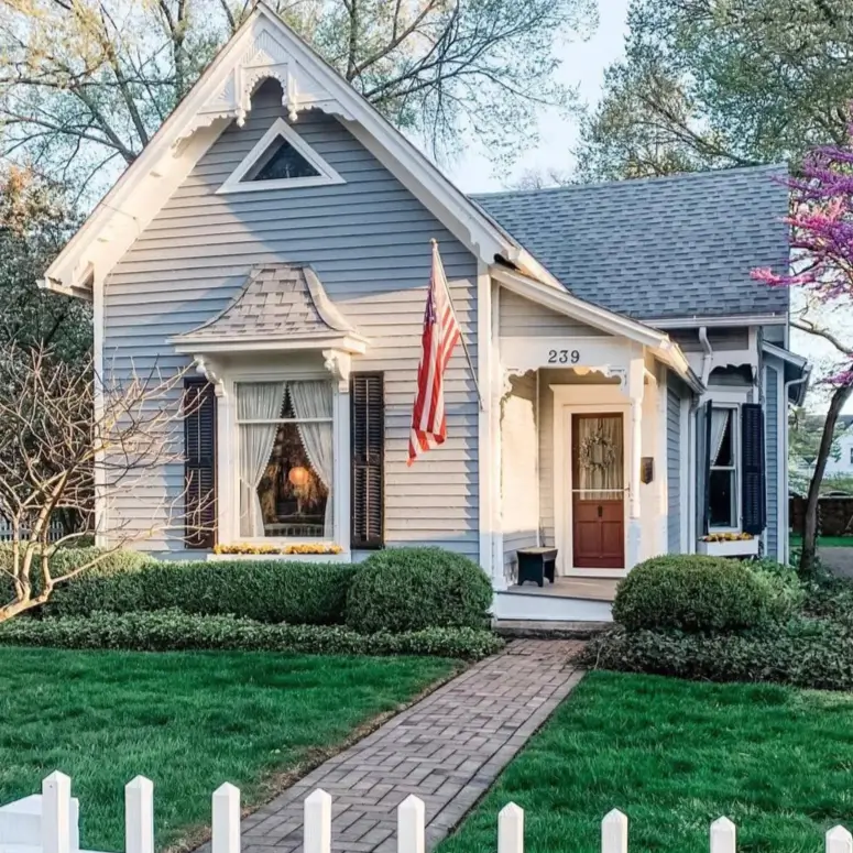 A charming light blue cottage with ornate white trim on the gable, a small bay window, and a white picket fence.