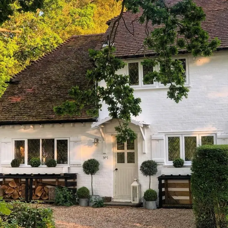 An English-style white brick cottage with a weathered tile roof and diamond-paned windows, set in a lush garden.