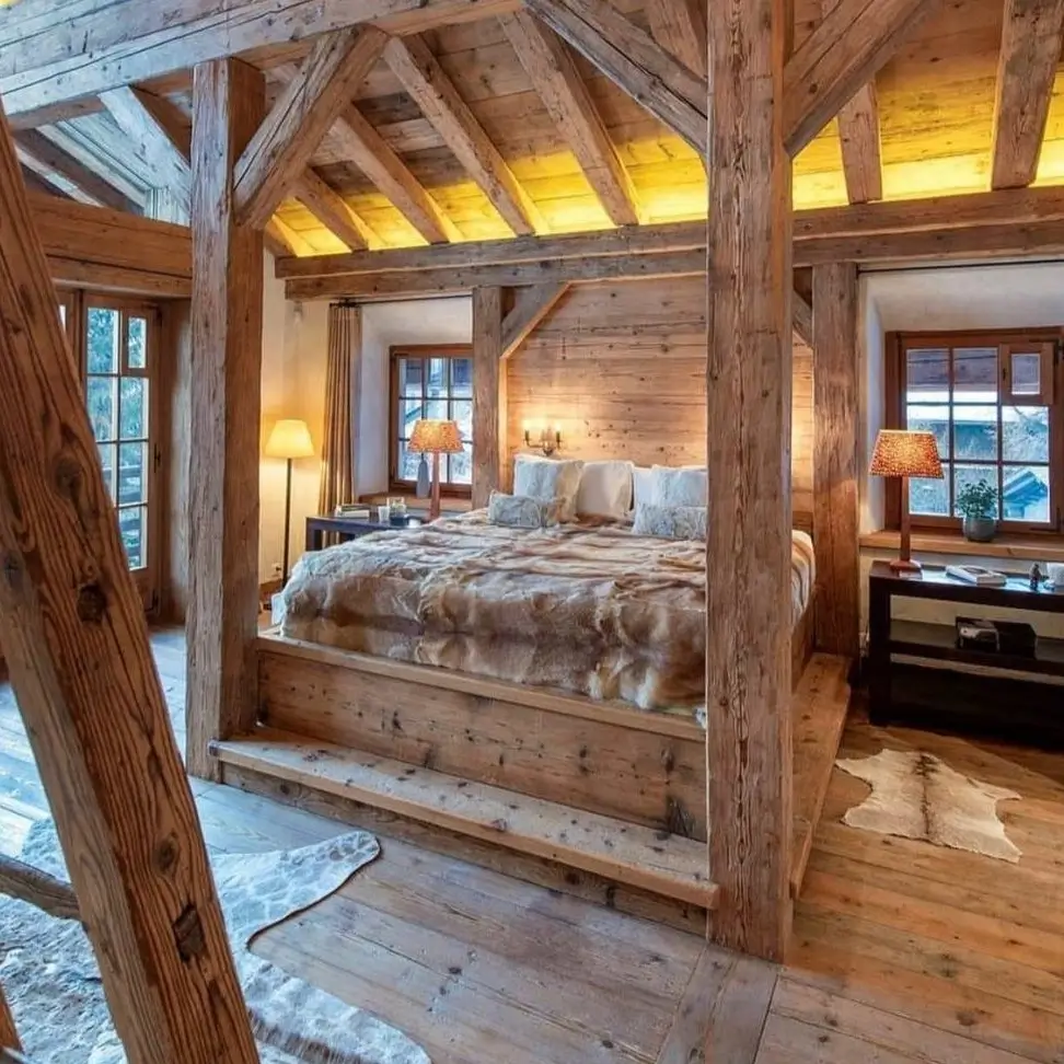 A rustic bedroom for couples featuring a large, architectural timber bed frame on a raised platform, covered with a fur blanket and illuminated by warm ceiling lights.