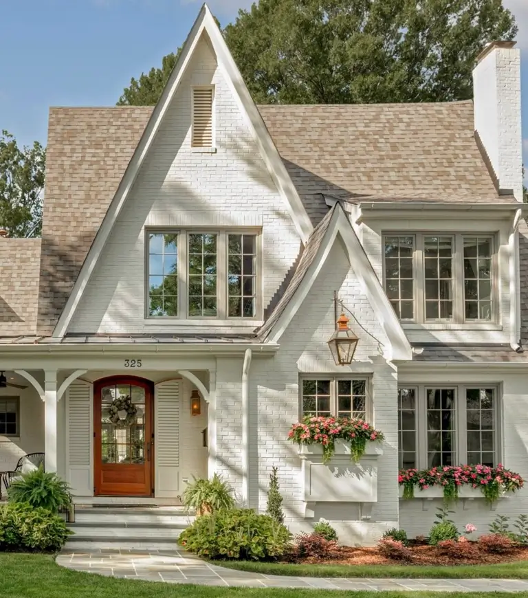 A white painted brick cottage with steep gables, a wooden front door, and window boxes filled with pink flowers.