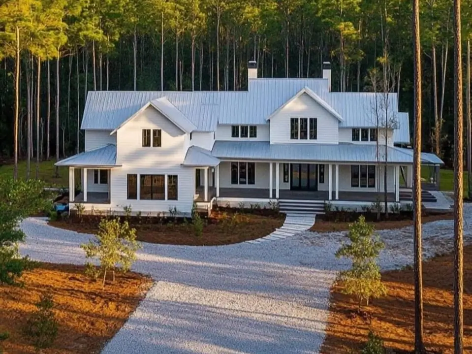 A sprawling modern white farmhouse with a light metal roof set in a tall pine forest with a gravel driveway.