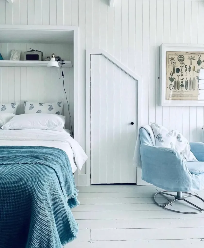 A bright white beach cottage bedroom with vertical wall paneling, a built-in bed, and a cozy blue armchair.