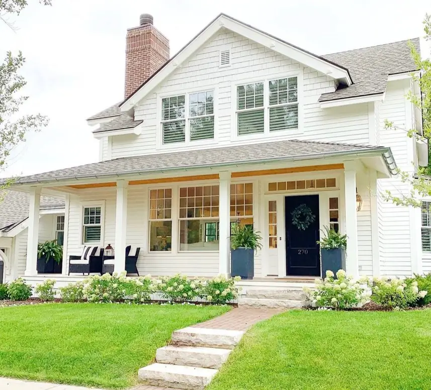 A white cottage with a large wraparound porch, a prominent brick chimney, and a dark front door flanked by planters.