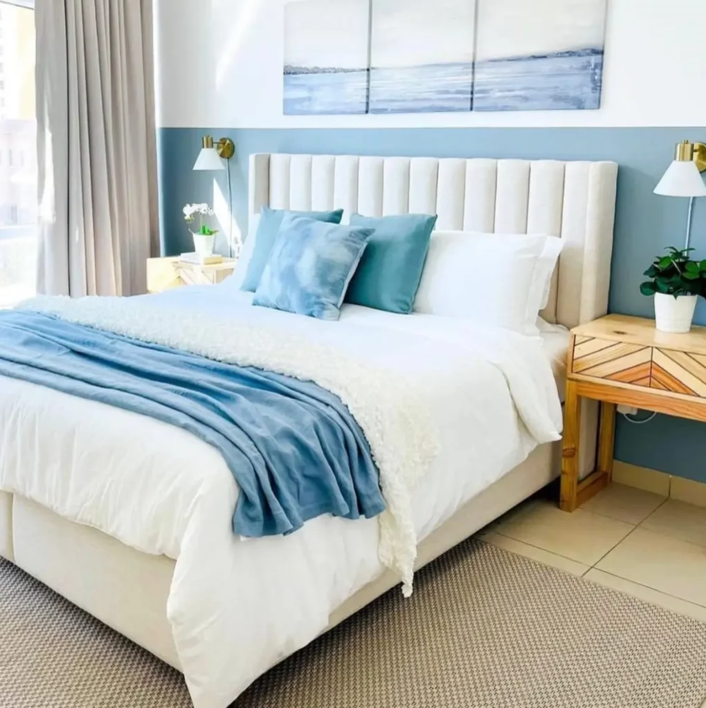 A beach theme bedroom with a two-tone blue and white wall, an upholstered bed, and abstract ocean art.