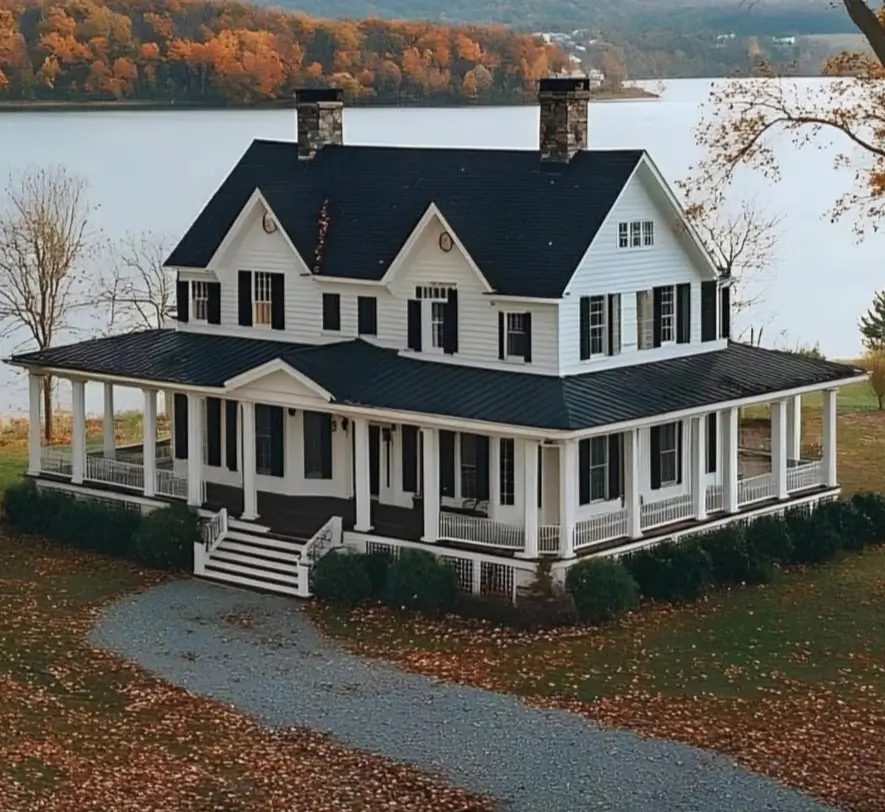 A stately white farmhouse with black shutters and a wraparound porch, set against a lake and autumn hills.