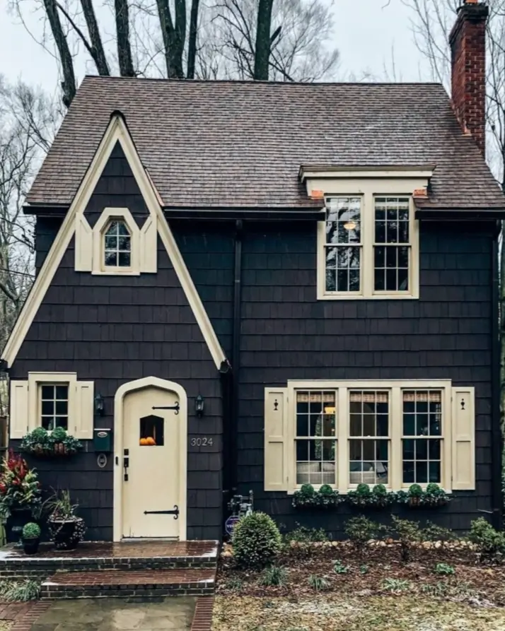 A storybook cottage with dark shingle siding, cream-colored trim and shutters, and an arched front door.