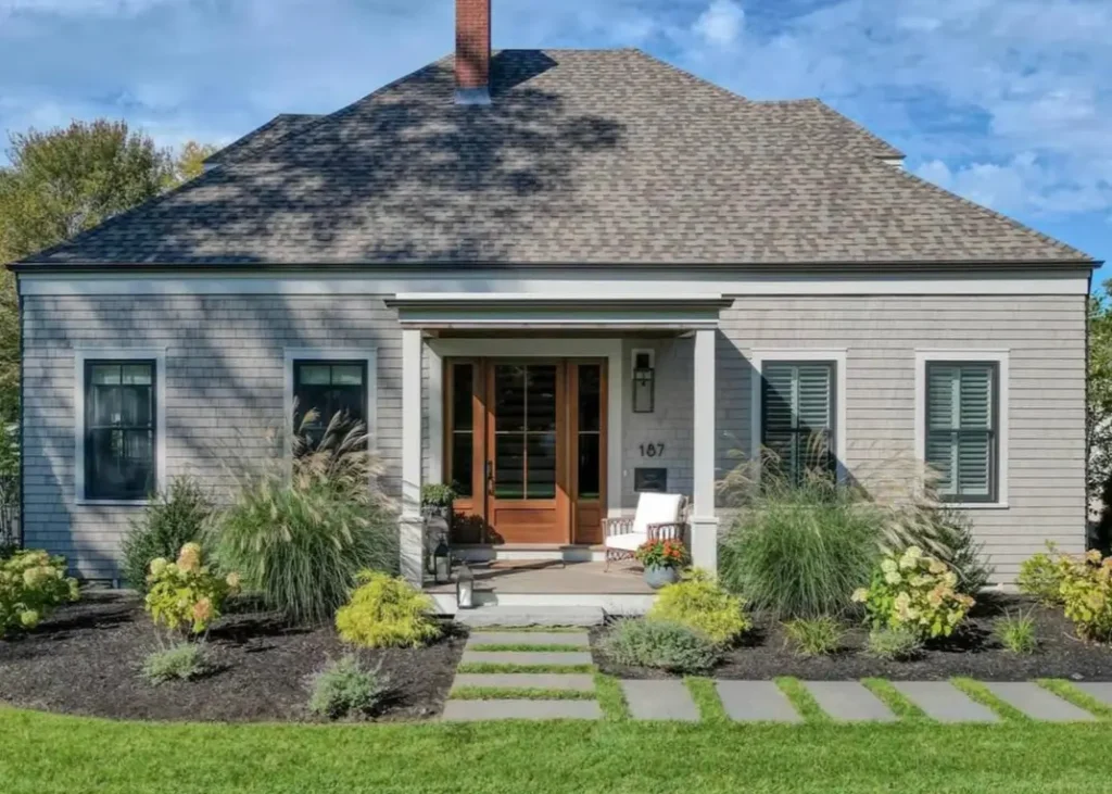 A modern single-story cottage with gray shingle siding, a wood front door, and large ornamental grasses in the front garden.
