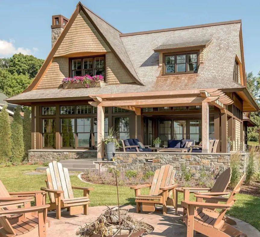 A cedar shingle cottage with a stone patio in the backyard, featuring a pergola and a fire pit with Adirondack chairs.