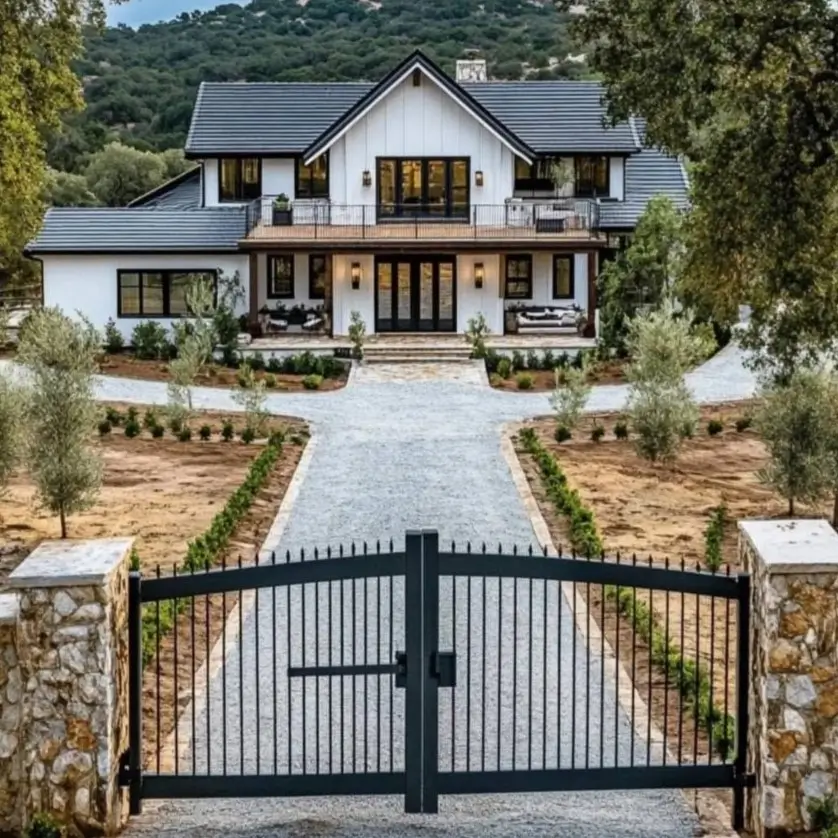 A modern white farmhouse with a second-story balcony, viewed through a black metal gate with stone pillars.