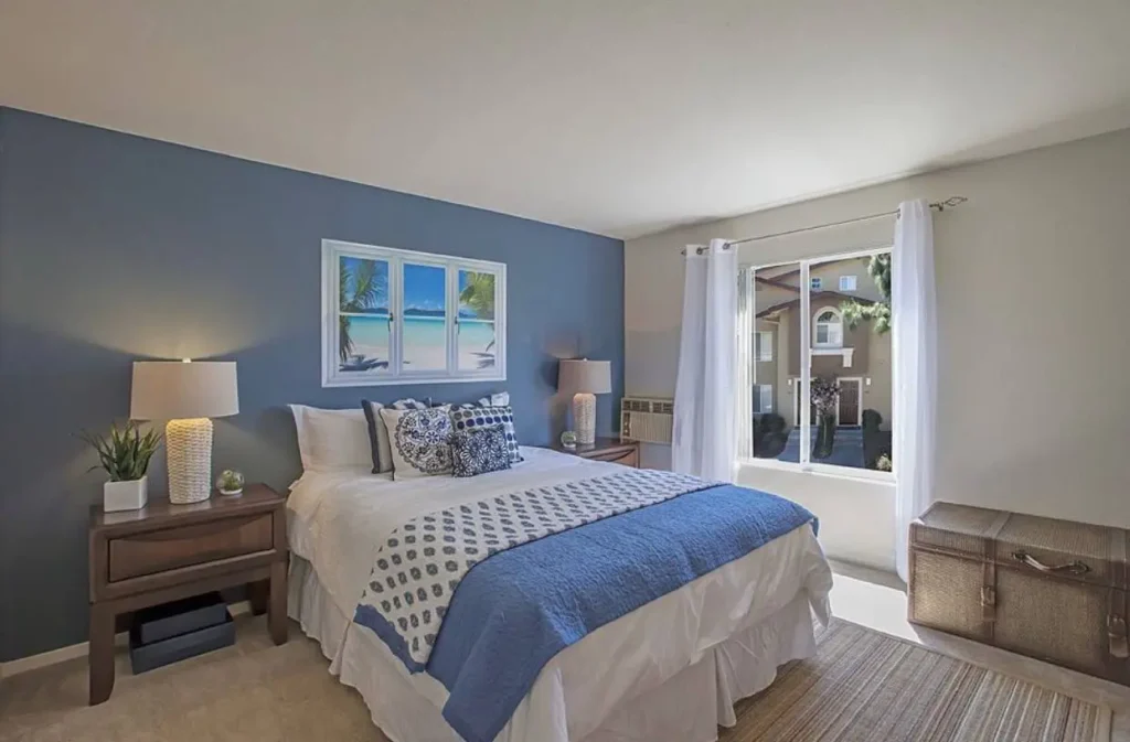 A bedroom with a blue accent wall featuring a piece of art depicting a sunny beach scene above the bed.