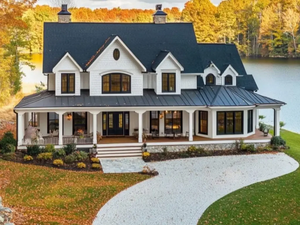 An elegant white farmhouse with a dark roof and a curved porch featuring large windows, overlooking a lake in autumn.