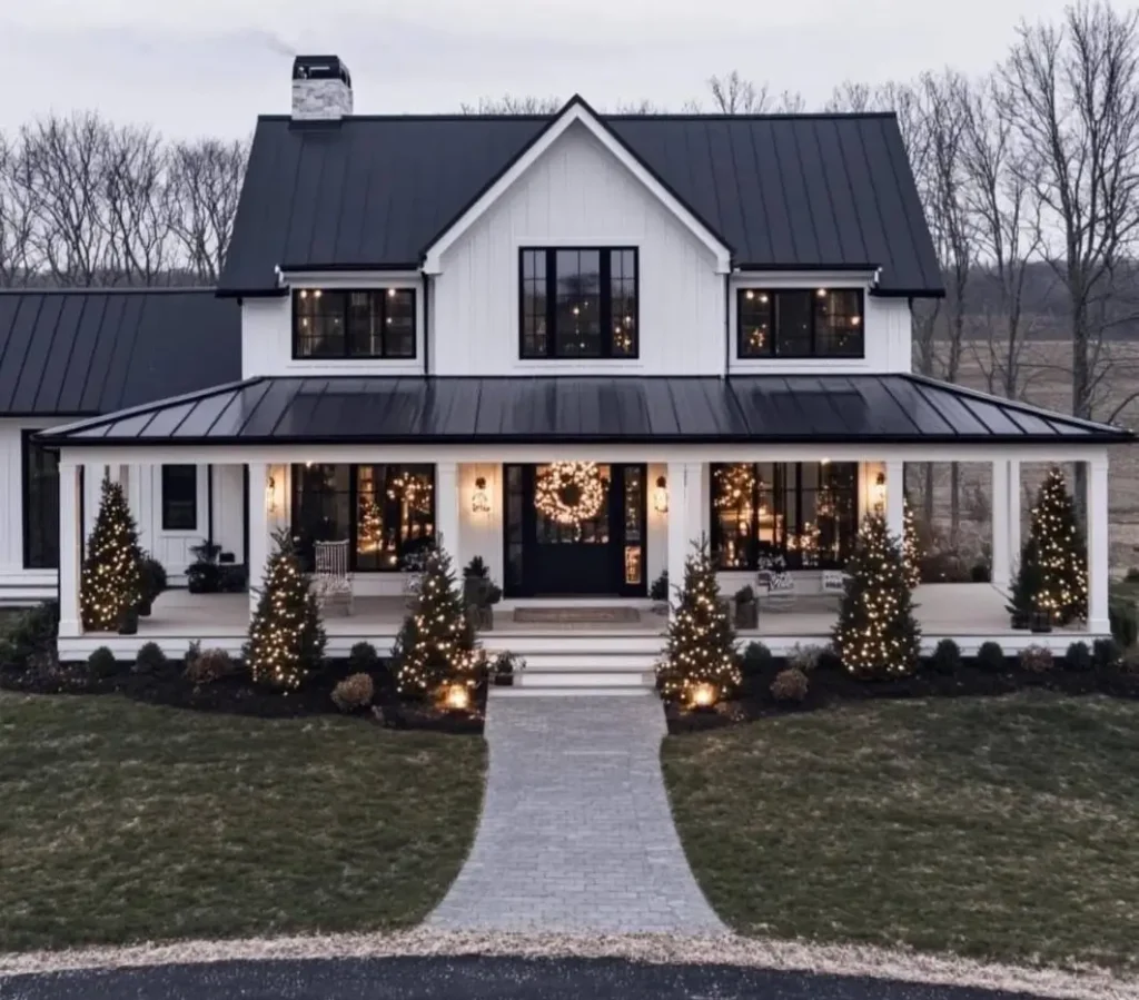 A modern white farmhouse decorated for Christmas with a large wreath and lighted trees on the porch at dusk.