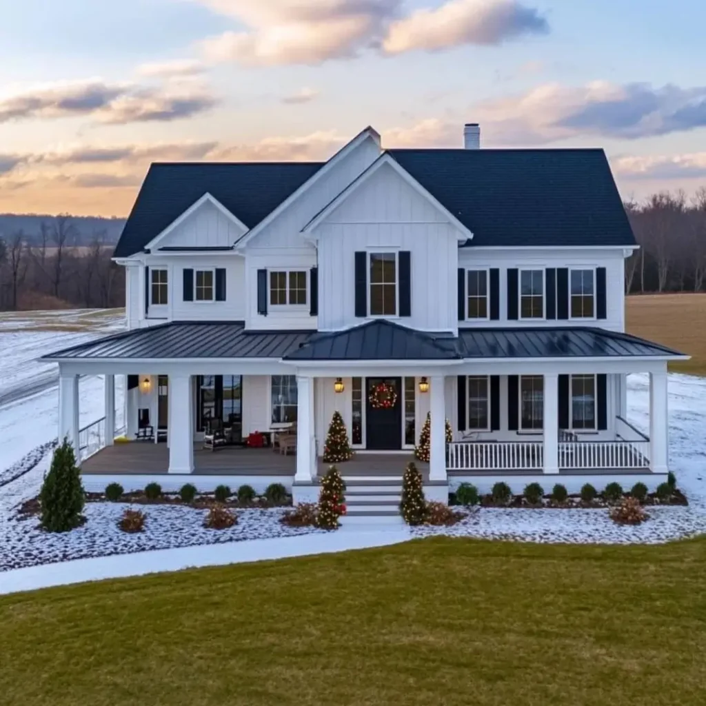 A modern white farmhouse with black shutters and simple Christmas decorations, set in a field with a light layer of snow.