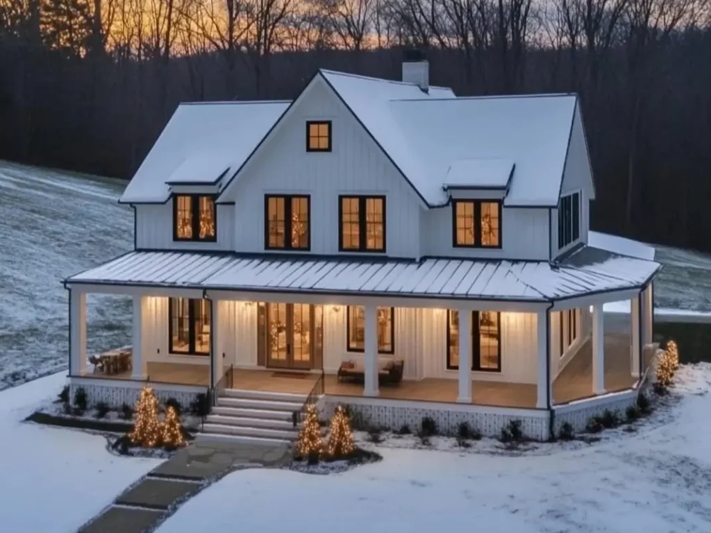 A snow-covered modern white farmhouse with warm lights glowing from the windows at sunset.