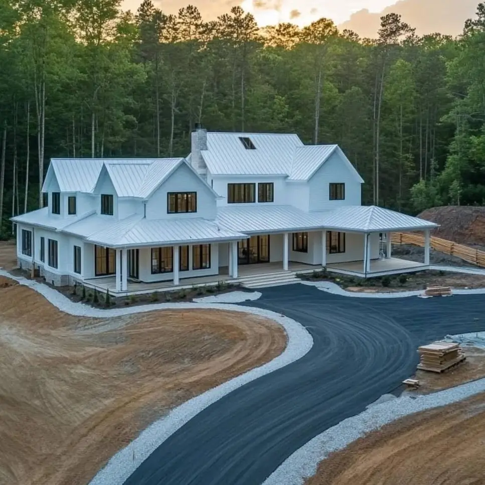 A large modern white farmhouse with a matching white metal roof and black windows, nestled in a wooded area.