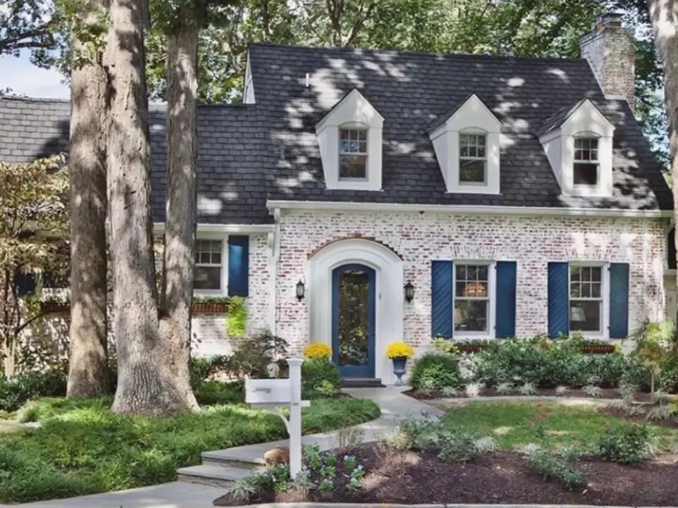 A white painted brick cottage with bright blue shutters and a matching front door, surrounded by mature trees.