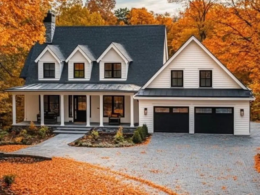 A modern white farmhouse with a trio of dormers and black garage doors, surrounded by bright orange autumn leaves.