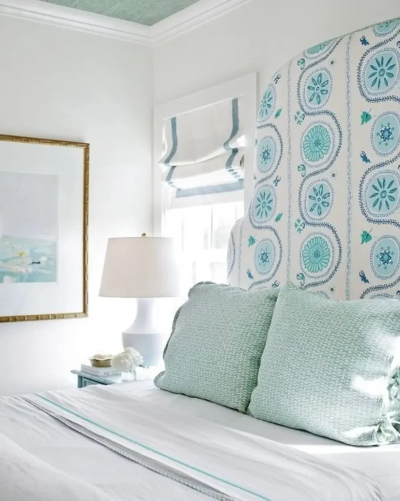 A bedroom with a tall upholstered headboard featuring a blue and aqua coastal pattern against a white wall.