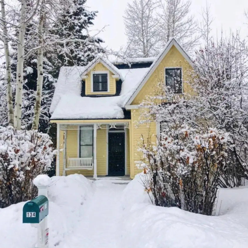 A small yellow cottage with a dark front door and a snow-covered roof and yard.