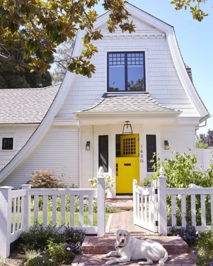 A white cottage with a gambrel roof and a bright yellow front door, with a white picket fence in the foreground.