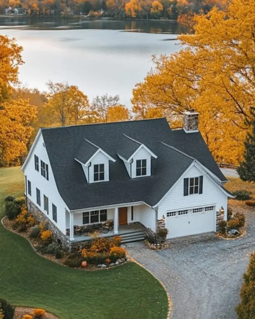 A white farmhouse with twin dormer windows and stone accents, surrounded by golden autumn trees near a lake.