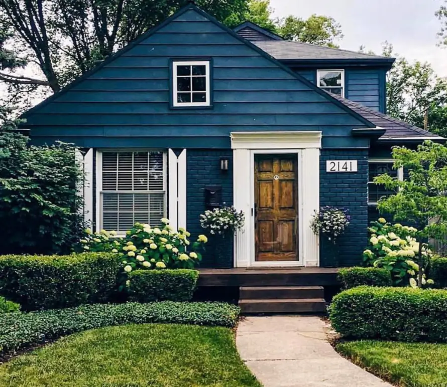 A deep blue cottage exterior featuring a natural wood front door, white trim, and framed by green hedges and hydrangeas.