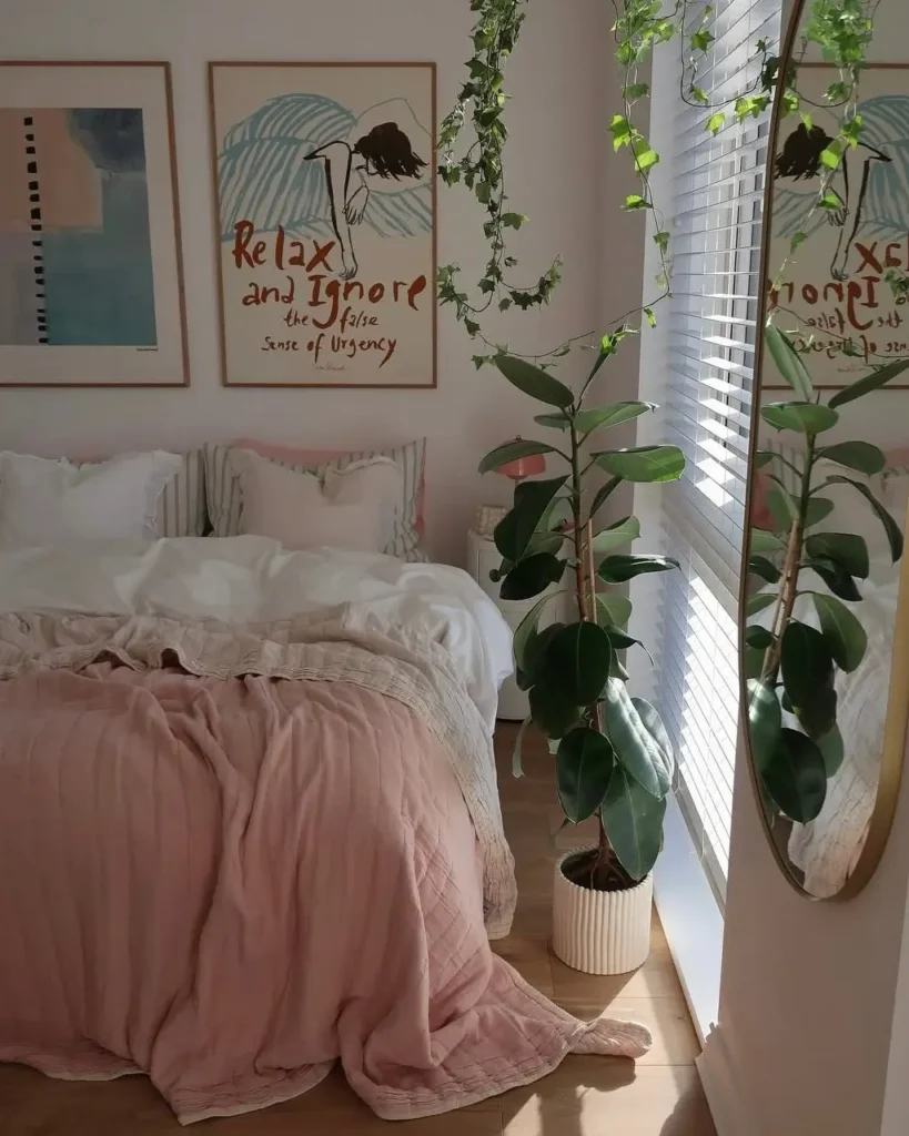 A bright bedroom with dusty pink bedding, a large rubber plant, and an oval mirror reflecting the window.