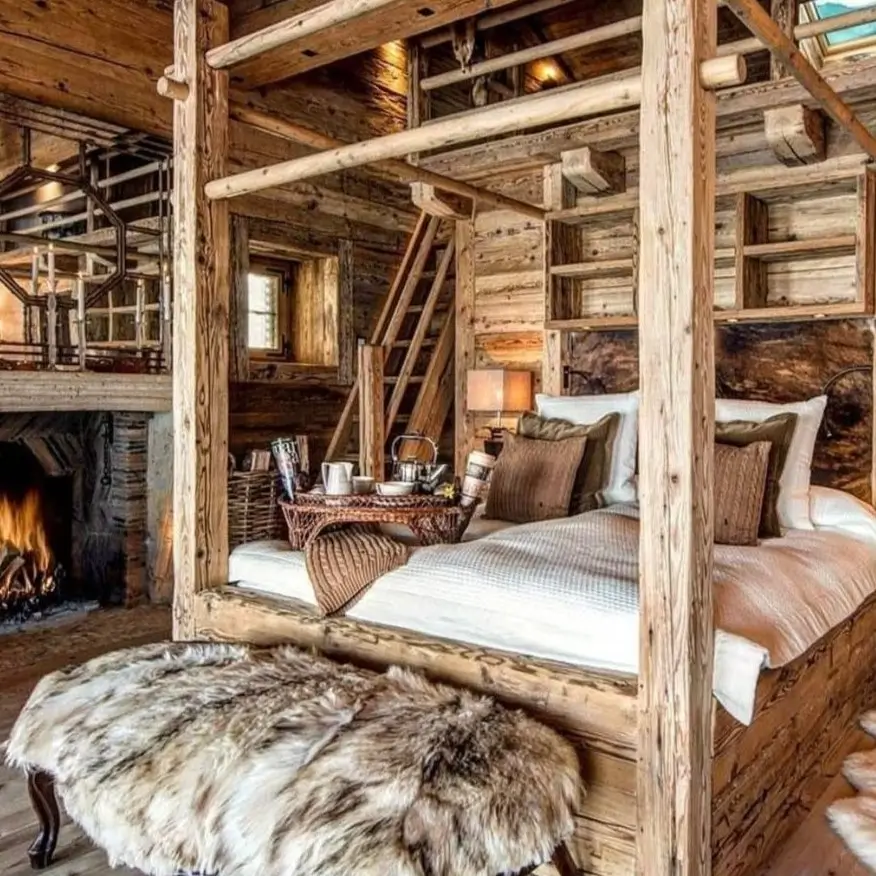 A cozy rustic bedroom for couples featuring a large wooden four-poster bed, a stone fireplace, and an elegant crystal chandelier hanging from a wood-paneled ceiling.