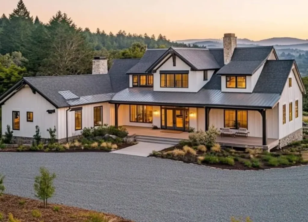 A sprawling modern white farmhouse with stone chimneys and dark trim, set against rolling hills at sunset.