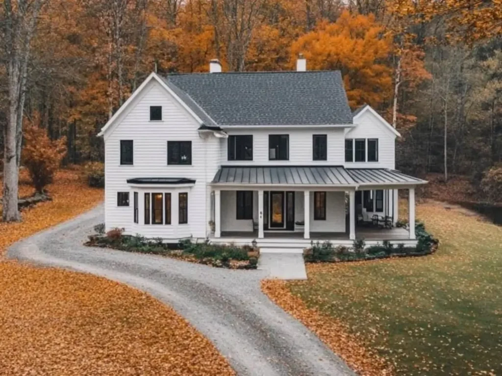 A modern white farmhouse with a bay window, nestled in a forest with vibrant orange autumn leaves.