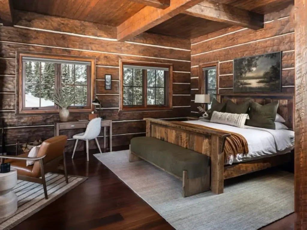A rustic log cabin bedroom for couples, featuring dark wood floors, a modern wooden bed with olive green pillows, and large windows with a winter view.
