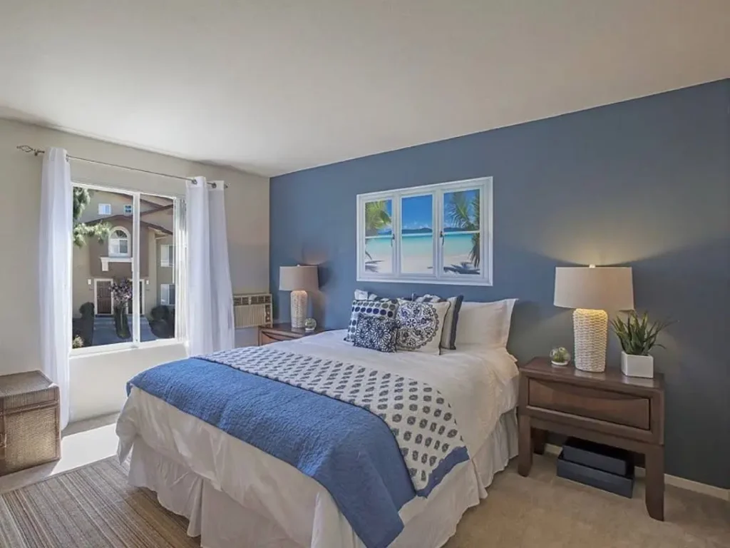 A beach themed bedroom featuring a blue accent wall, white and blue bedding, and a piece of art above the bed that looks like a window opening onto a tropical beach.
