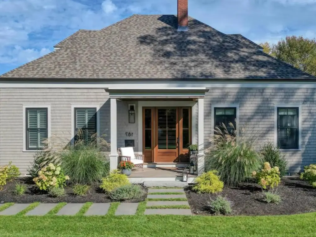 A modern cottage exterior with gray shingle siding, a natural wood front door, and a beautifully landscaped yard, showcasing charming cottage exterior ideas.