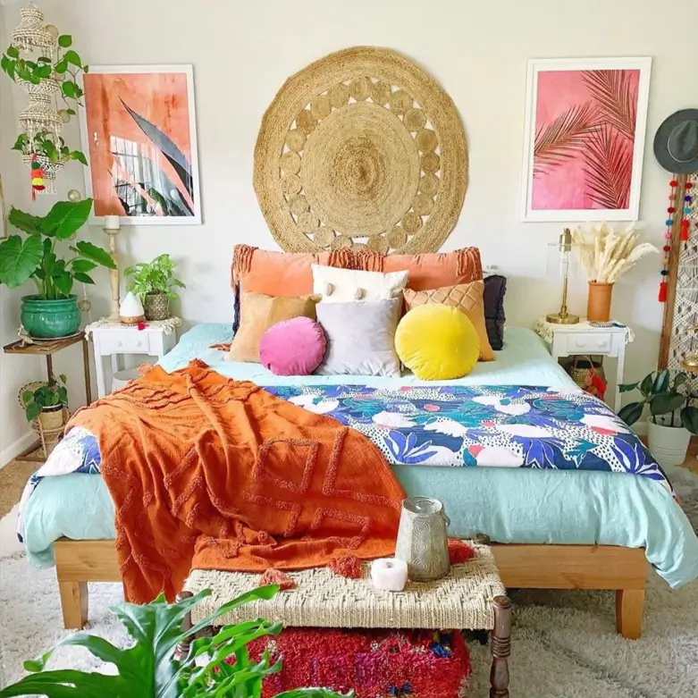 A colorful boho bedroom with a large round woven jute wall hanging centered above the bed, flanked by two pink botanical art prints.