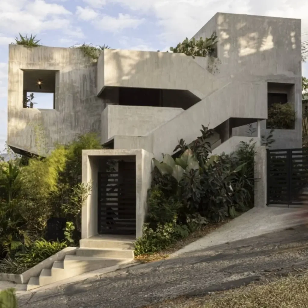 The striking concrete exterior of a brutalist house, its clean lines softened by lush greenery cascading from the roof and an external staircase.