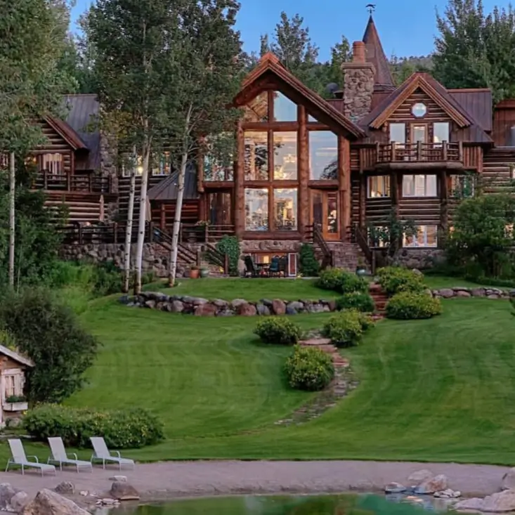 Embracing Grandeur: 25 Inspiring Ideas for Large Log Cabin Homes 4 A large log cabin home featuring a central glass wall, stone chimneys, and a multi-level design nestled in a wooded area.