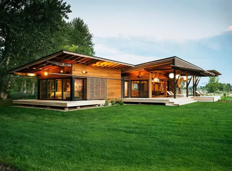 A modern ranch house exterior with natural wood siding and a large wraparound deck under an evening sky.