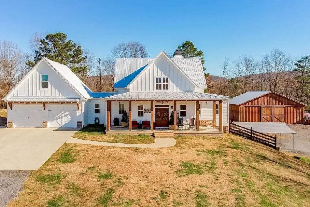 A modern white farmhouse exterior featuring a long front porch with natural wood columns, a metal roof, and a detached garage, set against a backdrop of rolling hills.