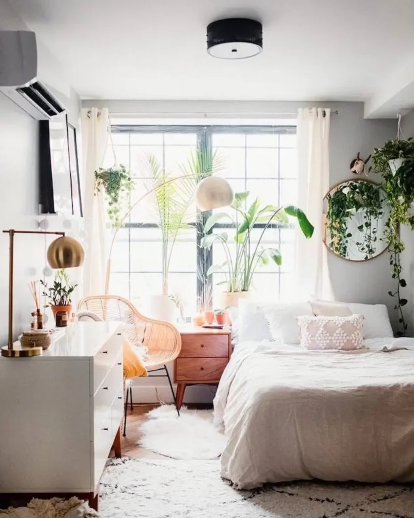 A small cozy bedroom with a large window, white bedding, a wicker chair, and numerous green houseplants creating a bright and airy design.