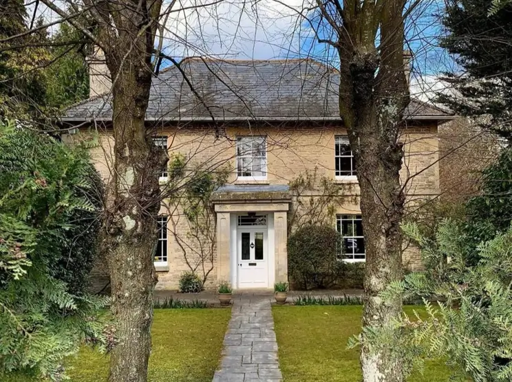 A symmetrical British country house with a stone exterior and white front door, framed by two large trees.