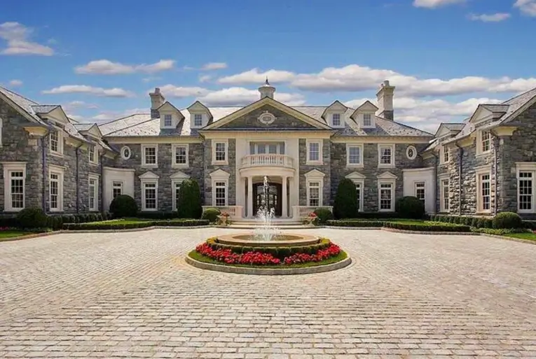 20 Stone Mansion Ideas for Classic & Modern Designs 12 A massive, symmetrical stone mansion with a vast cobblestone motor court and a central fountain with red flowers.